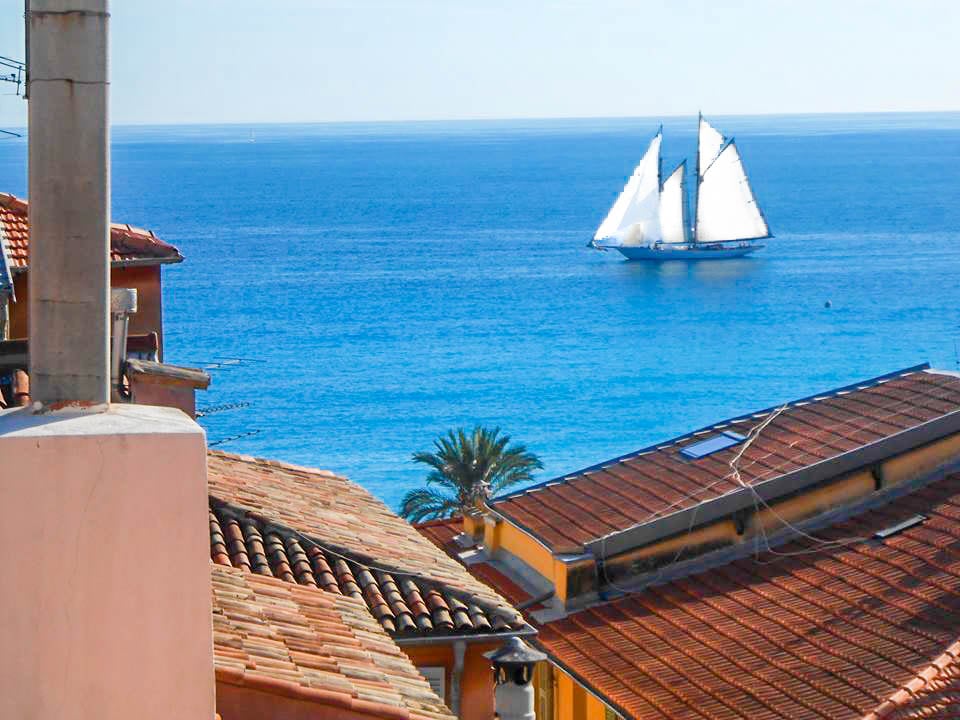 A sweeping view of the sea is presented, with a sailboat gracefully navigating the calm waters. The rooftops of Old Nice are visible in the foreground, featuring traditional Mediterranean tiles. Palm trees are partially seen, enhancing the coastal scene under clear blue skies.