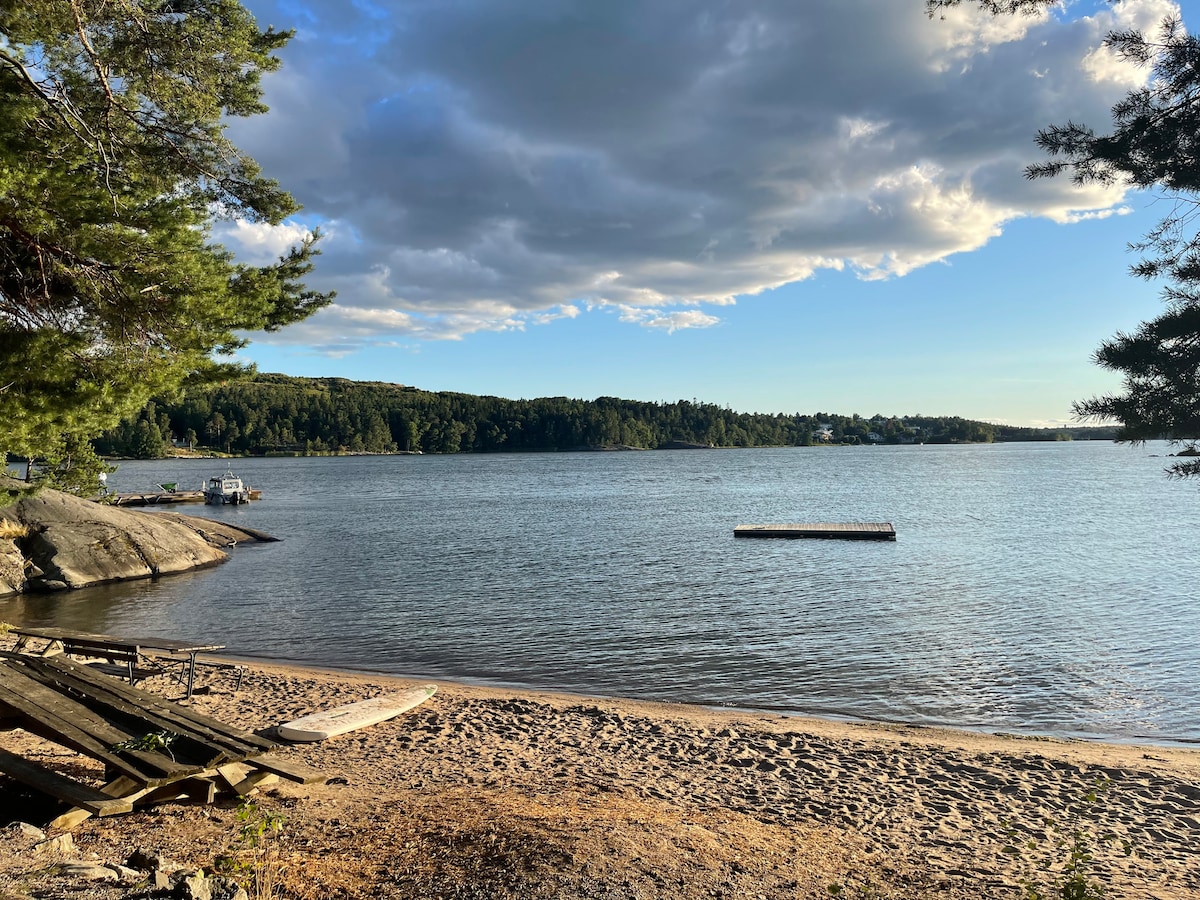 A serene view of a sandy beach with calm waters extending to the horizon, surrounded by lush greenery. A wooden dock is visible in the distance, while several boats are moored nearby. Soft clouds drift overhead, enhancing the tranquil atmosphere of the island.