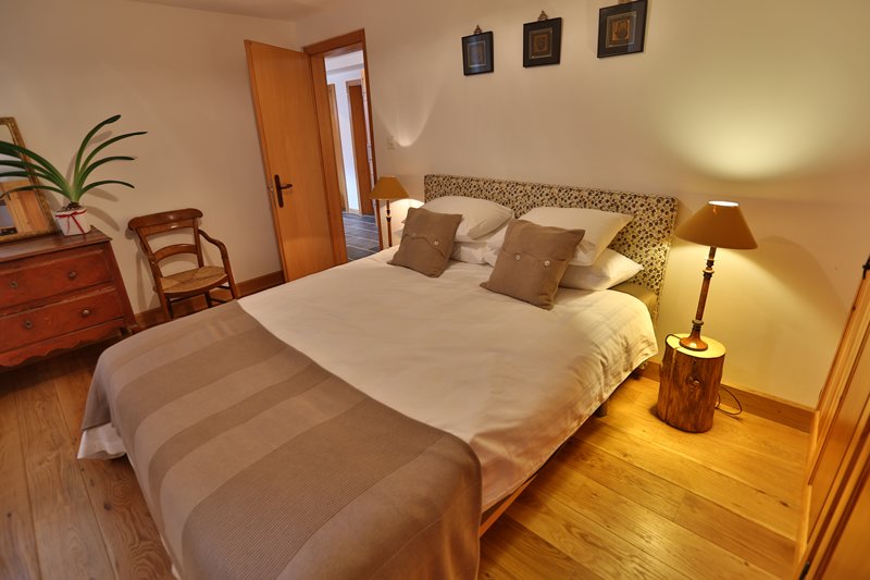 A spacious bedroom features a large bed with decorative pillows and a neutral-colored blanket. Two bedside lamps provide soft lighting, and a wooden chair is placed to one side. The room showcases natural wood flooring and framed artwork on the walls.