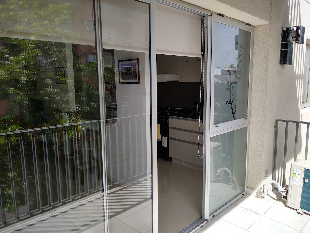Sliding glass doors open to a spacious balcony, allowing natural light to brighten the area. A modern kitchen is visible through the doors, featuring sleek cabinetry and contemporary appliances. The balcony's railing is designed with horizontal bars, offering a clear view of greenery outside.