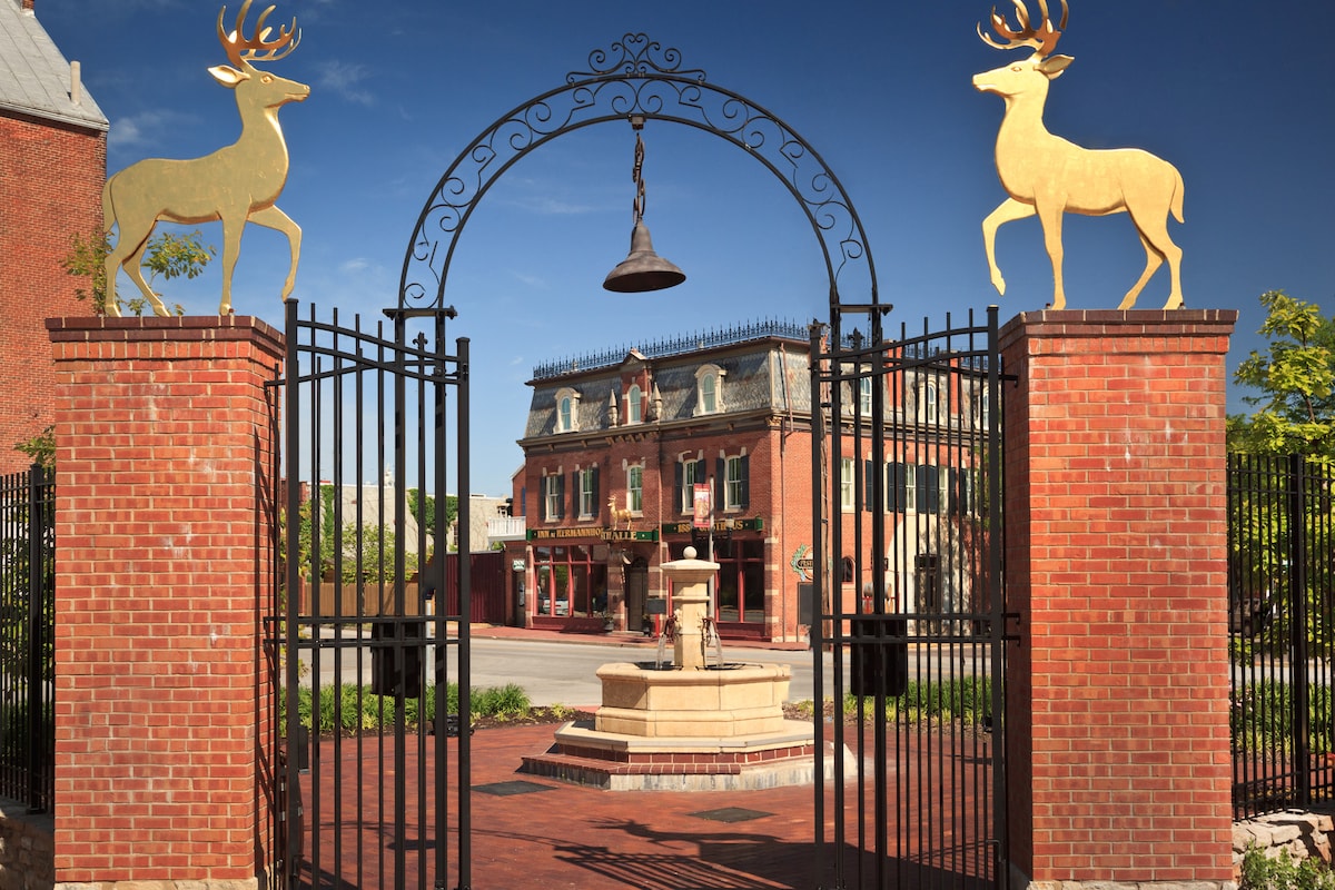 An elegant entrance is framed by two golden deer sculptures atop brick columns. A bell hangs centrally above wrought iron gates, leading to a picturesque courtyard with a fountain. The historic building can be seen in the background, showcasing classic architecture against a clear blue sky.