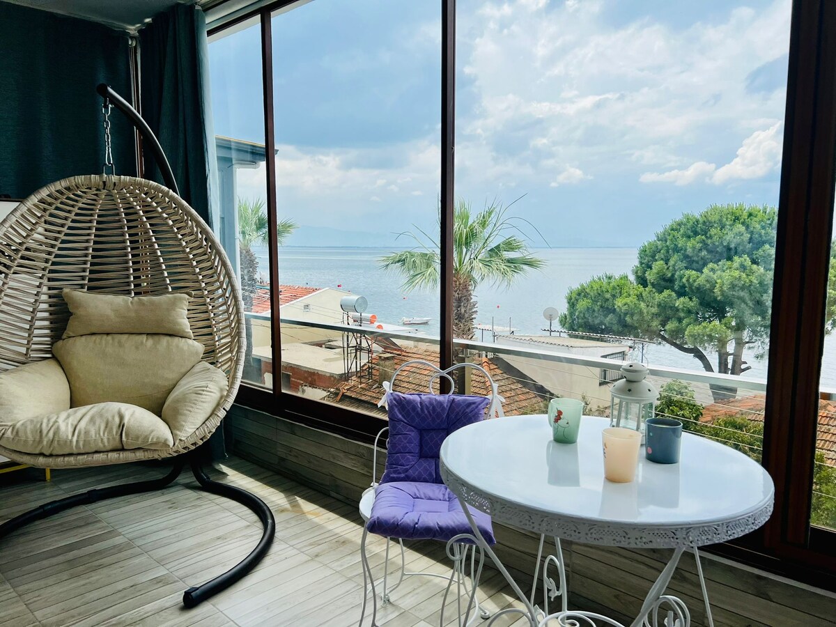 A cozy seating area features a woven hanging chair and a small circular table. Natural light fills the space through large windows, providing a view of the serene ocean, palm trees, and a peaceful outdoor terrace.