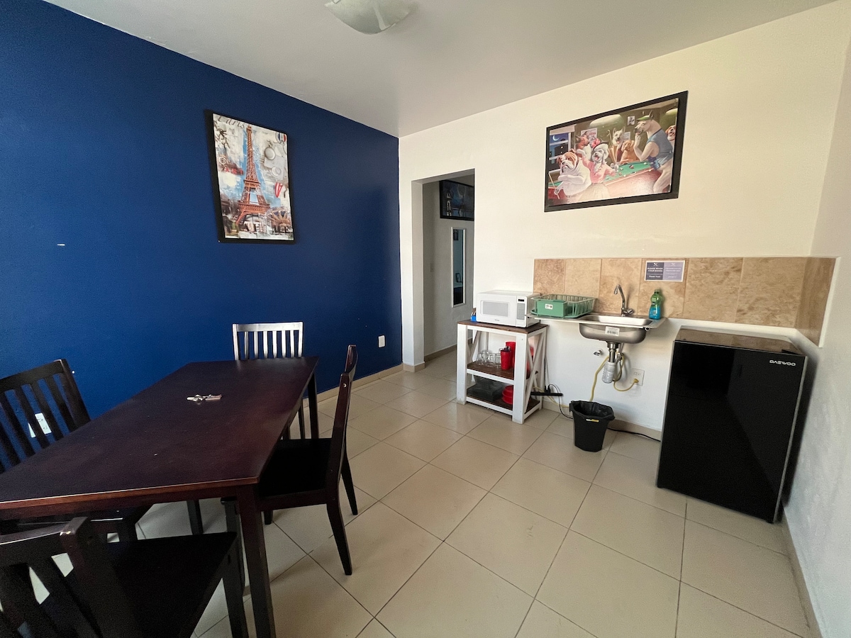 A dining area features a dark wooden table surrounded by four chairs, set against a vibrant blue accent wall. A framed artwork adds character above the table. A compact kitchenette is visible, equipped with a sink, microwave, and a small refrigerator.