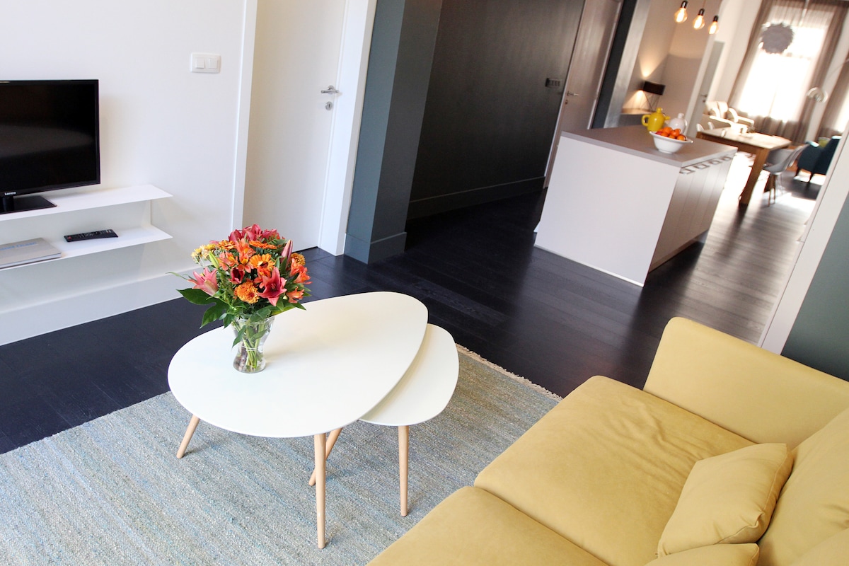 A bright living area features a yellow sofa and a white coffee table with a floral arrangement. Soft lighting enhances the space, with a view towards an open kitchen and dining area in the background. Dark hardwood floors complement the modern decor.