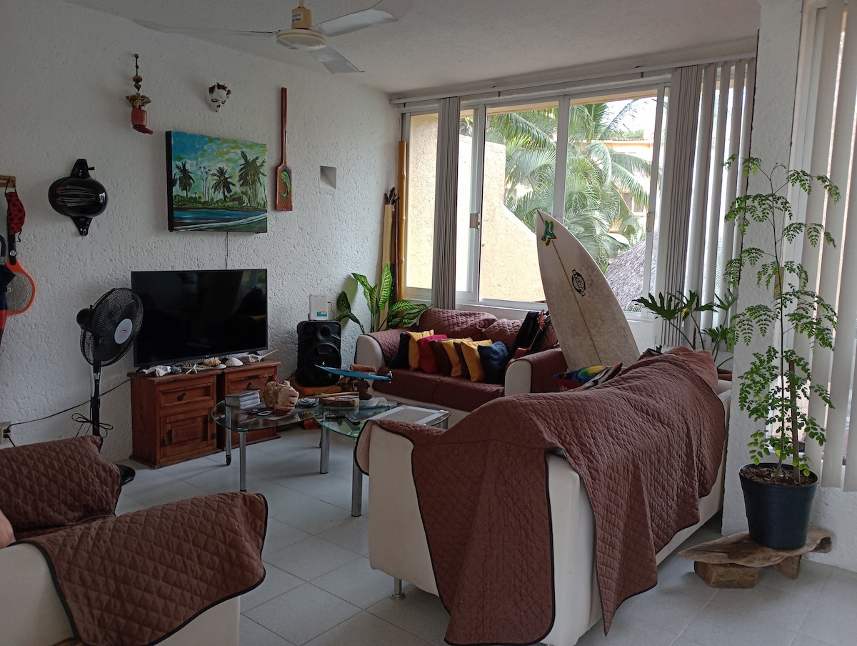 A bright living area features large windows that invite natural light, complemented by tropical plants. Comfortable seating is provided on sofas covered with brown quilts. A surfboard leans against the wall, and a television is positioned on a wooden cabinet, enhancing the casual atmosphere.