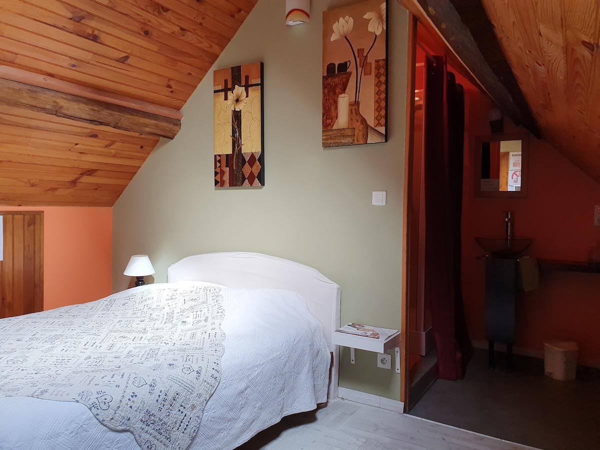 A cozy attic bedroom features a spacious bed topped with a patterned blanket. The walls are adorned with abstract art, and soft lighting is provided by a bedside lamp. A modern sink area is visible through an open doorway, enhancing the room's functionality.