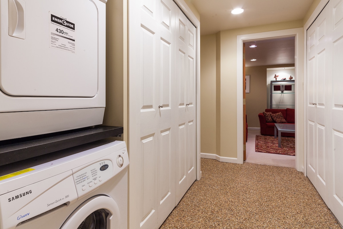 A laundry area features a stacked washer and dryer, positioned against a neutral-colored wall. Dual white doors provide storage space on the opposite side of the hallway. Soft lighting illuminates the space, with a glimpse of a cozy living area visible in the distance.
