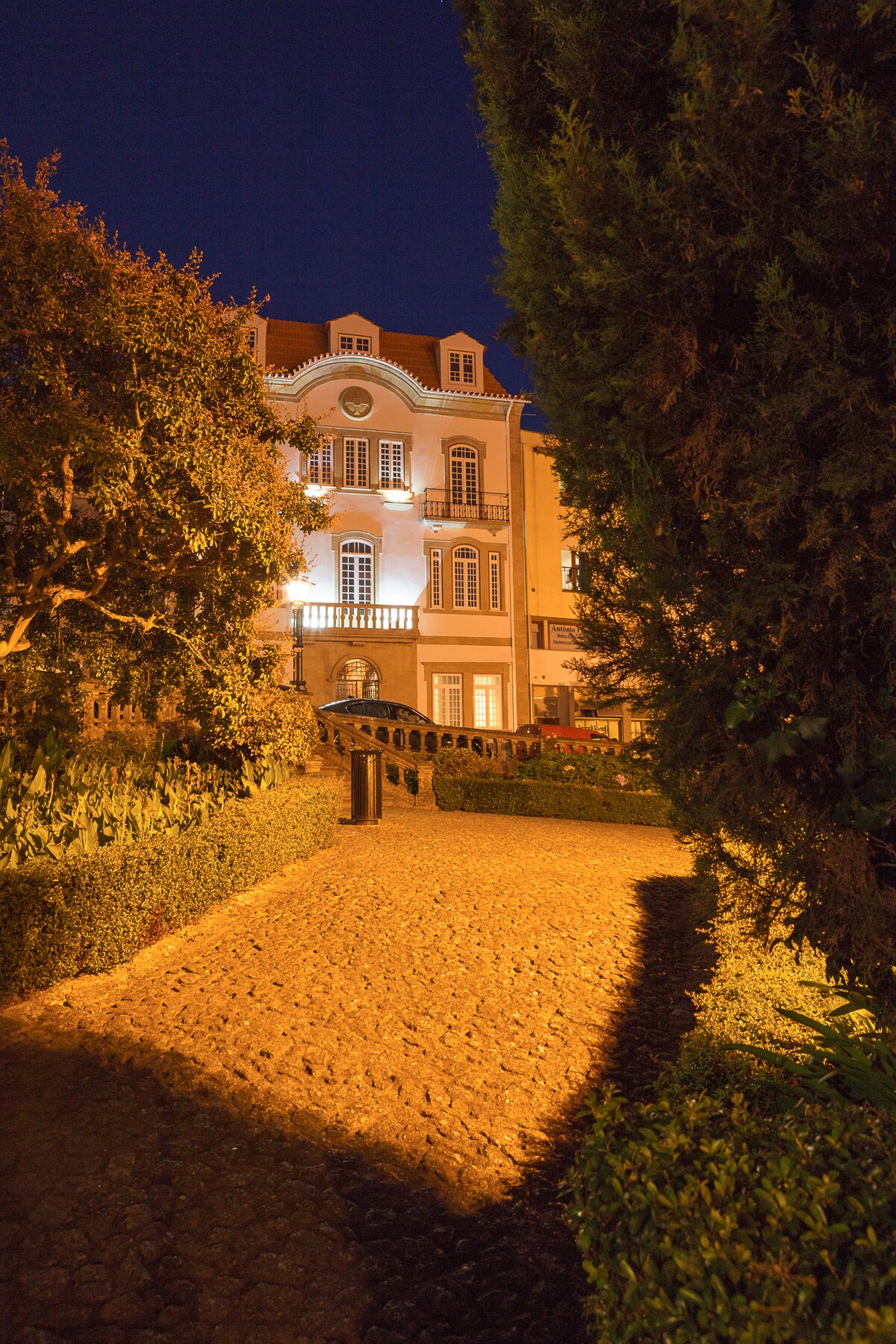 The pathway is lined with neatly trimmed hedges, leading to a beautifully illuminated building. Soft lighting highlights architectural features of the structure, while the surrounding greenery adds a serene touch to the scene.