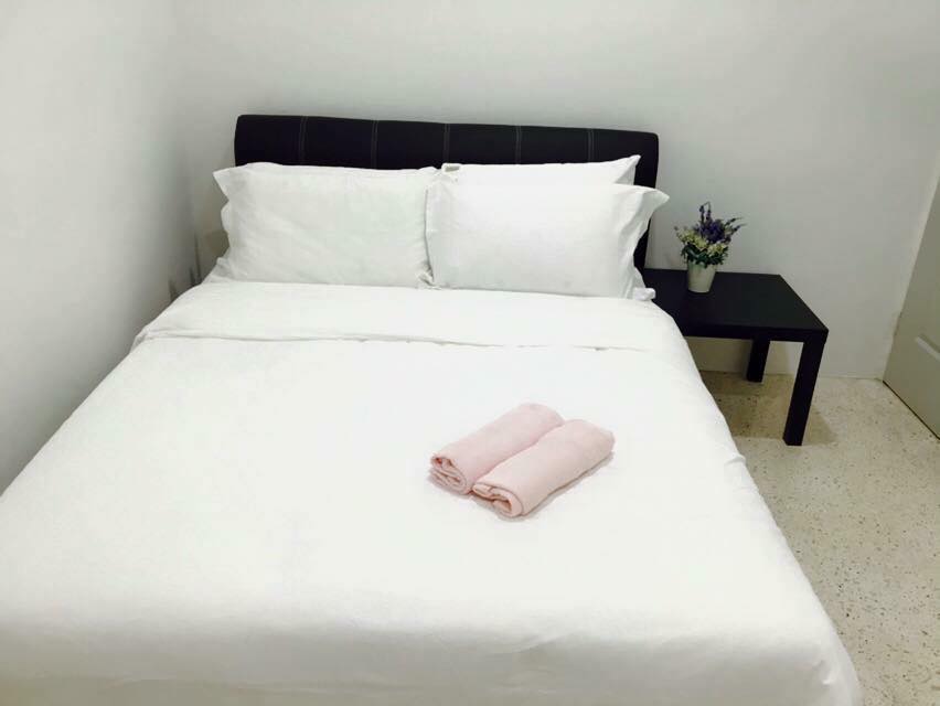 A neatly arranged sleeping area features a spacious bed with crisp white linens and plush pillows. A small bedside table holds a potted plant, adding a touch of greenery. Two rolled towels in soft pink are placed centrally on the bed.