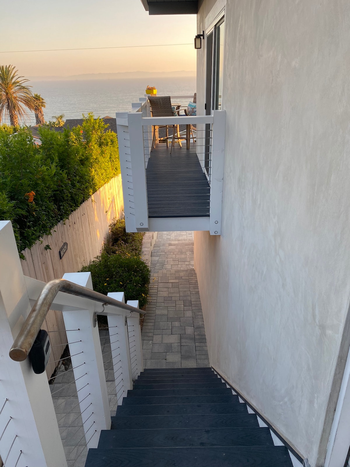 A staircase leads down to a path bordered by lush greenery, with a view of outdoor seating above. The boardwalk-style balcony extends from the house, connecting the two levels and offering glimpses of the ocean in the distance during sunset.