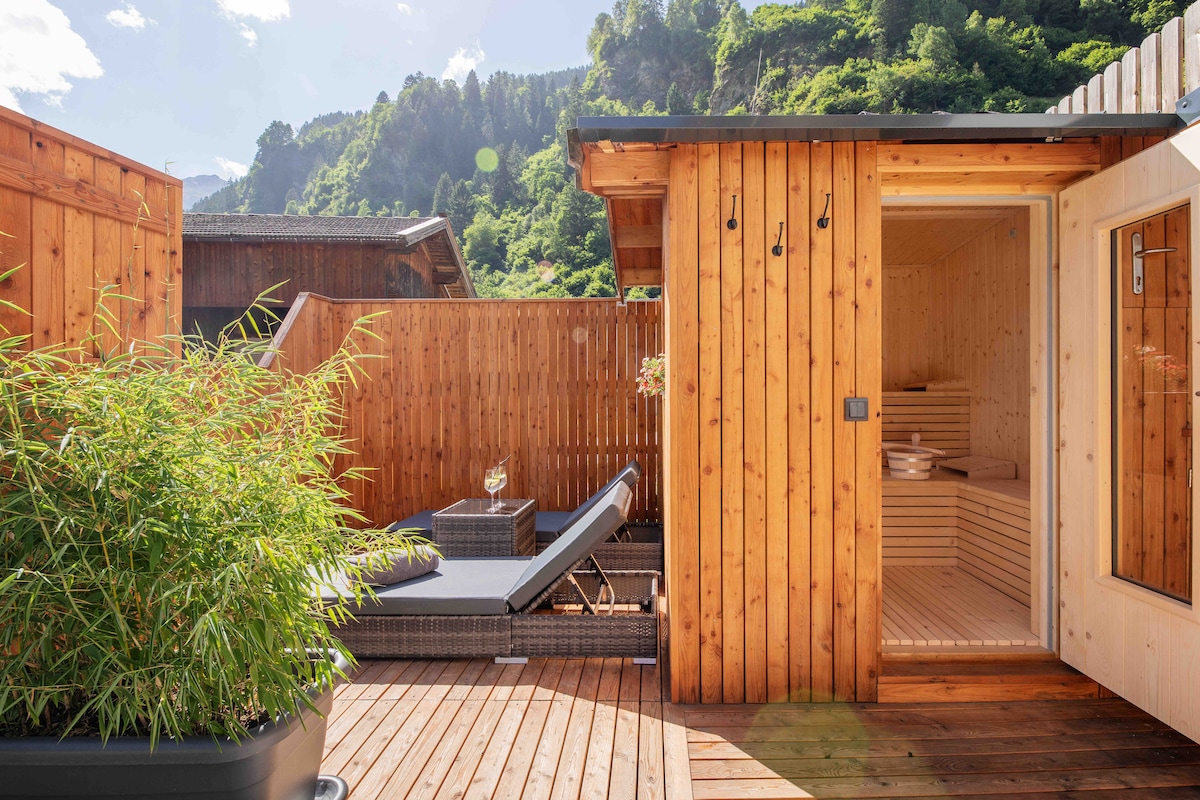 A wooden terrace is surrounded by lush greenery, featuring a sunbed and a small table with a glass. A sauna cabin with a glass door stands nearby, offering easy access to the outdoor space. The overall setting is bright and inviting.