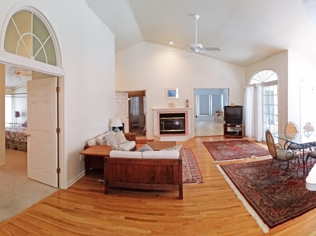 An open living space features high cathedral ceilings with two large windows allowing natural light. A wooden coffee table sits in front of a full couch and loveseat. A fireplace is centered on the wall, with a TV positioned nearby. Elegant rugs cover the hardwood floor.