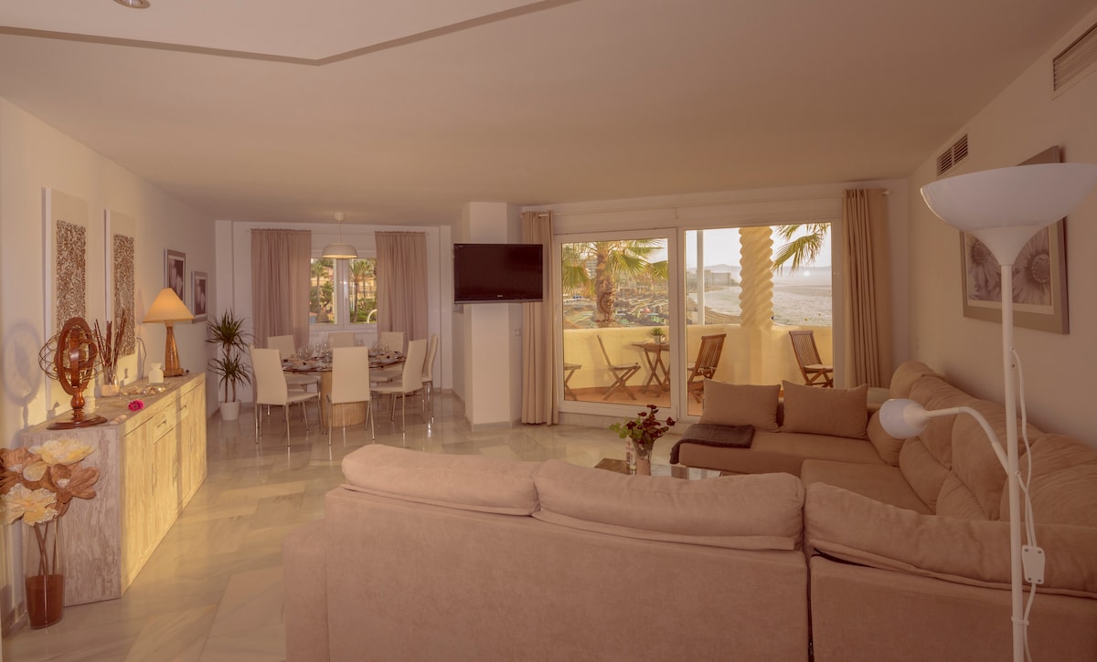 An open and airy living area features large glass doors that offer a view of the terrace and beach. A cozy sofa invites relaxation, while a dining table is set for gatherings. Natural light fills the space, enhancing the soft color palette of the furnishings.