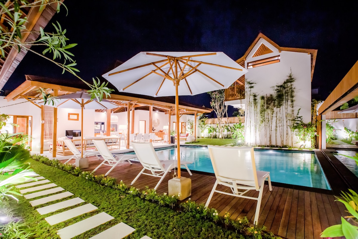 The outdoor area features a tranquil swimming pool surrounded by lush greenery and modern loungers. A large umbrella offers shade over the seating area, which includes a wooden table. Soft lighting enhances the serene atmosphere in this open space.