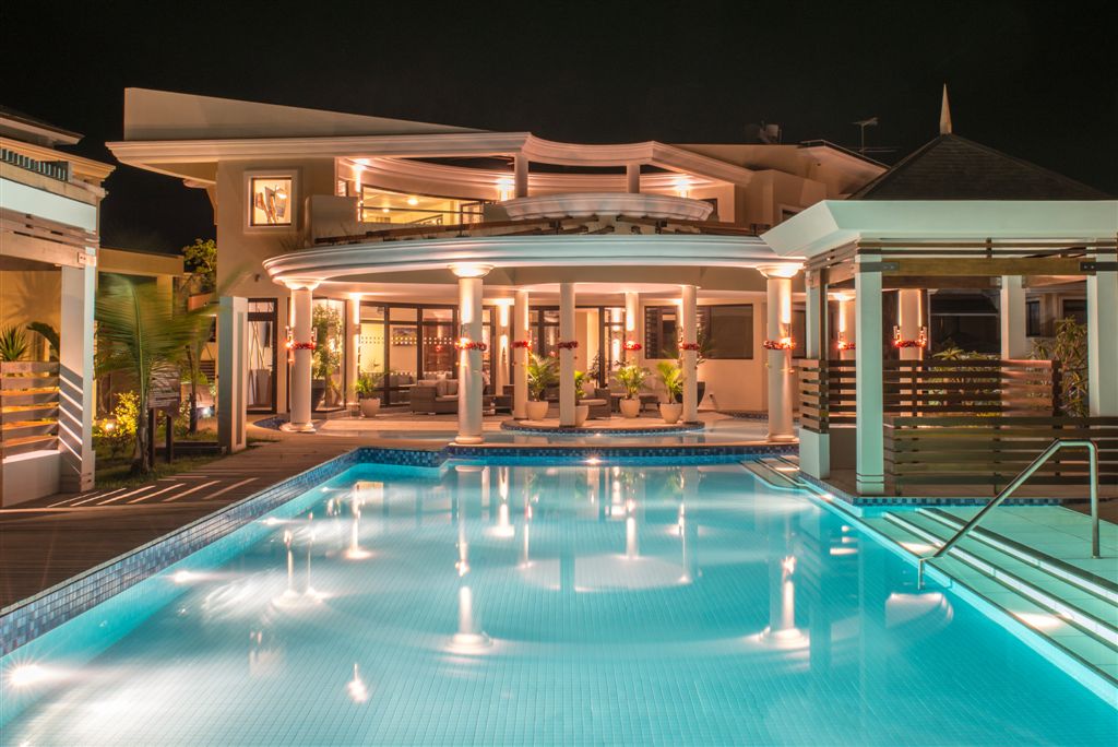 A modern villa is showcased at night, illuminated by warm lighting. The spacious outdoor area features a pristine swimming pool, surrounded by comfortable seating and lush greenery, inviting guests to relax and enjoy the tranquil ambiance.