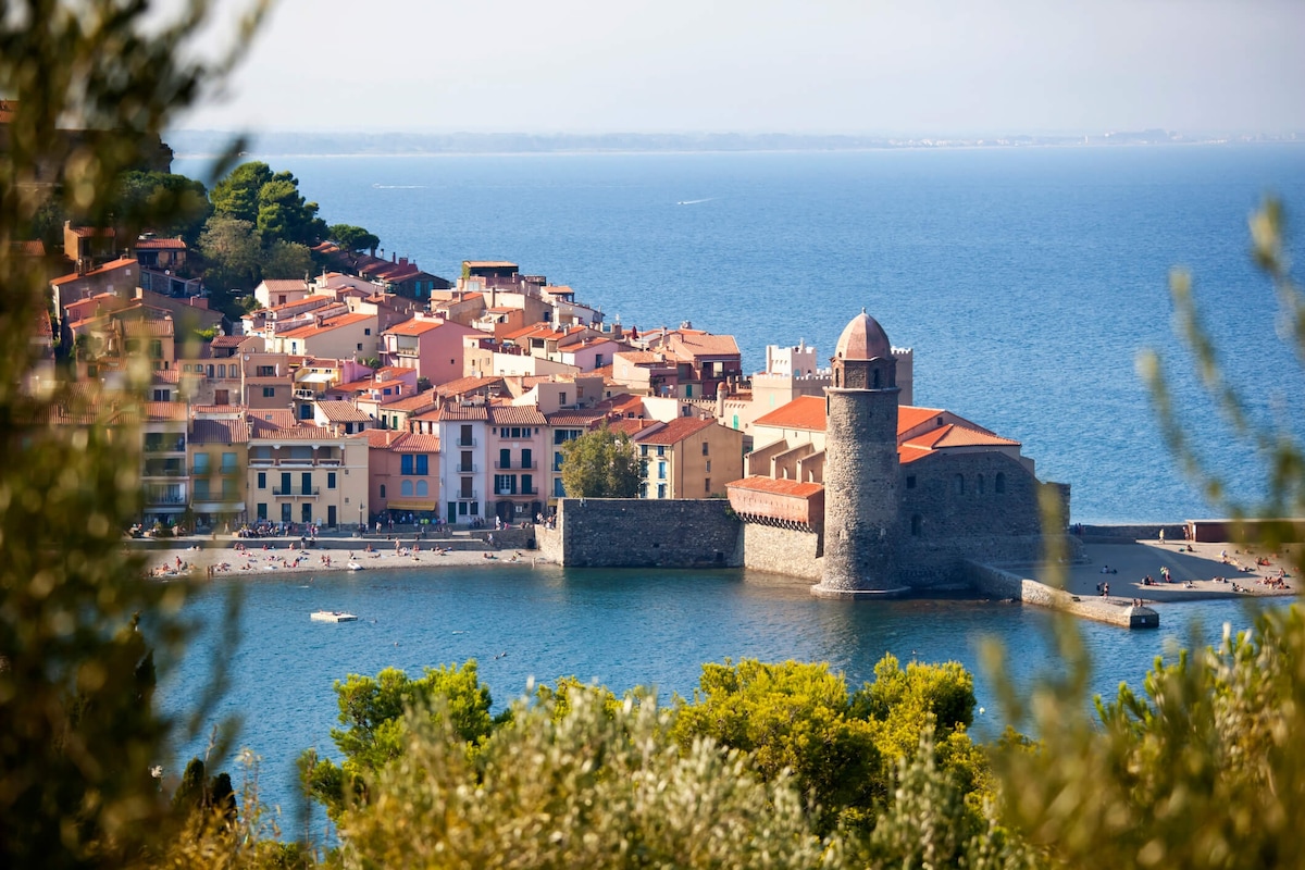 A panoramic view showcases the coastal town, featuring colorful buildings lining the shore. The foreground highlights the clear blue water, with a historical stone structure prominently positioned along the beach. Lush greenery surrounds the scene, enhancing its natural beauty.