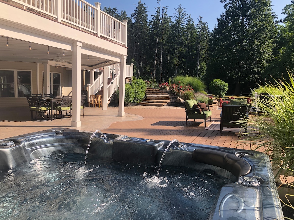 A spacious wooden deck surrounds a hot tub with water gently cascading over its edges. Comfortable outdoor seating is visible nearby, framed by lush greenery and trees in the background, creating a serene setting for relaxation.