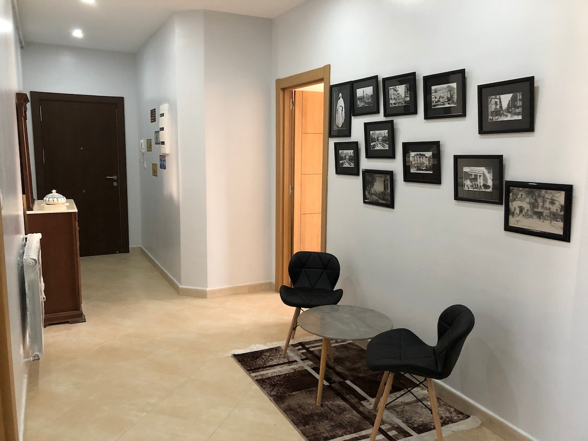 A hallway is shown, featuring a light-colored tiled floor. Two modern chairs are positioned around a circular table. Black and white framed photographs adorn the wall, adding character to the space. A wooden cabinet is visible in one corner, contributing to the room's overall ambiance.