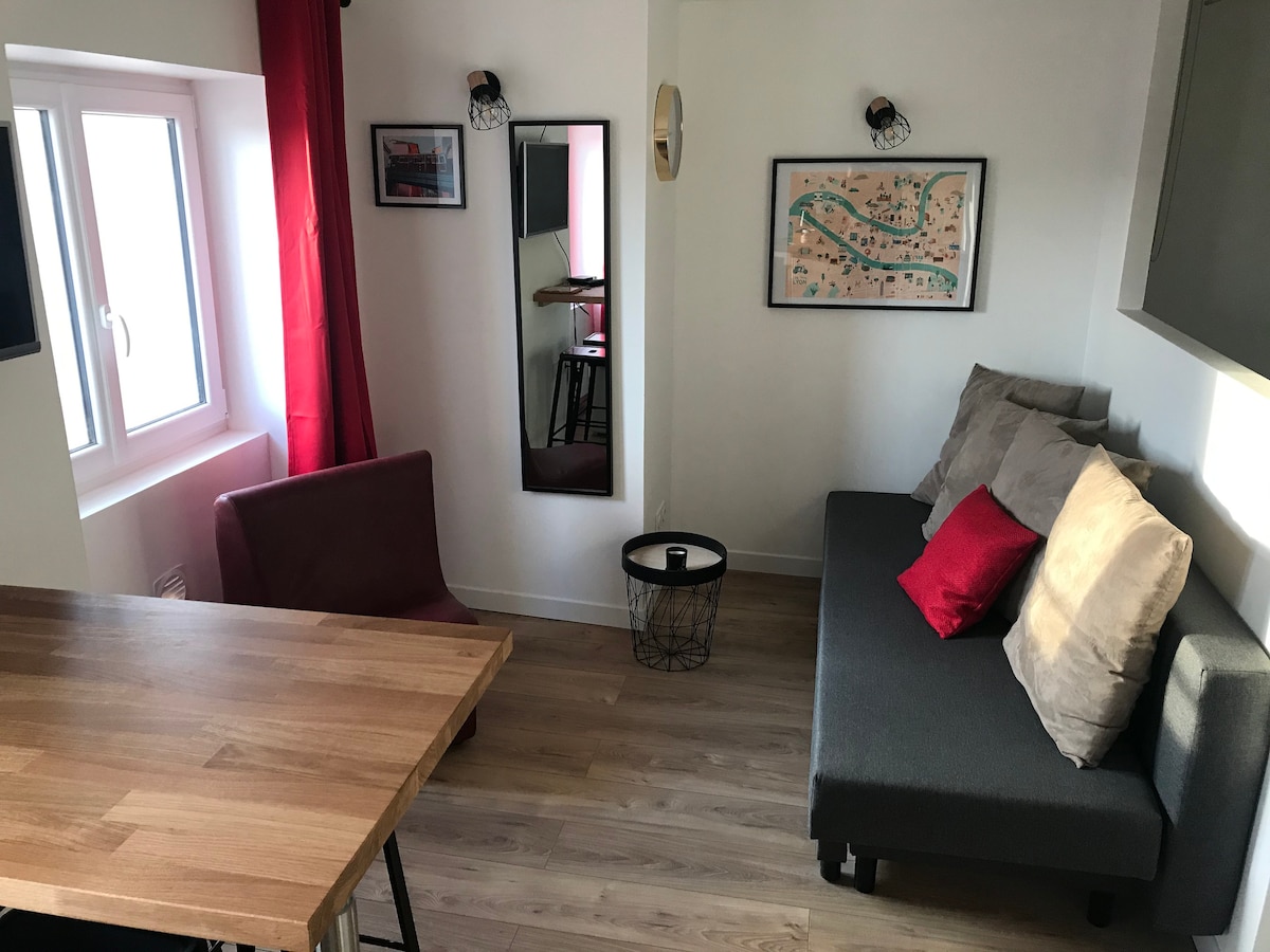 A compact living area is showcased, featuring a gray sofa with accent pillows and a wooden dining table. A red curtain flanks a window allowing natural light to brighten the space. Wall art and a modern light fixture are seen in the background, contributing to the ambiance.
