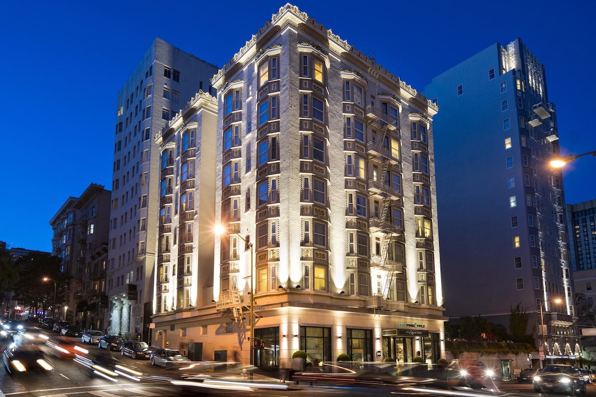 A historic hotel building is illuminated against the evening sky, showcasing its intricate architectural details and elegant façade. Surrounding streets are lined with vehicles, highlighting the vibrant urban environment of San Francisco's Union Square.