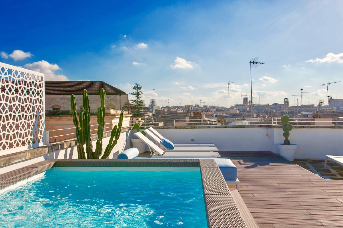 A spacious terrace features a hot tub surrounded by several sun loungers. Potted plants, including a tall cactus, add greenery to the area. The view overlooks a vibrant city skyline under a clear blue sky.