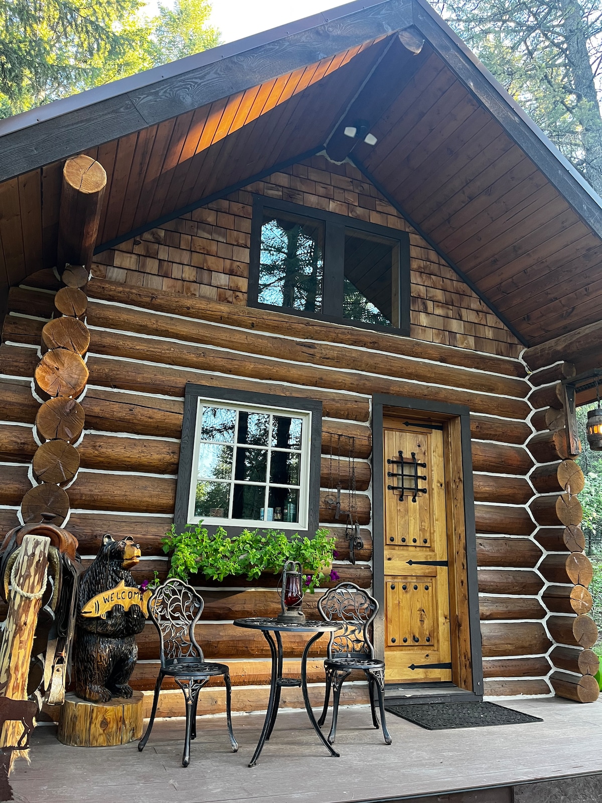 The cabin's entrance features a wooden door with a decorative grille, flanked by two windows. A small table with two ornate chairs is positioned on the porch, enhancing the welcoming appearance of the log construction. Planters with green foliage are visible beside the door.