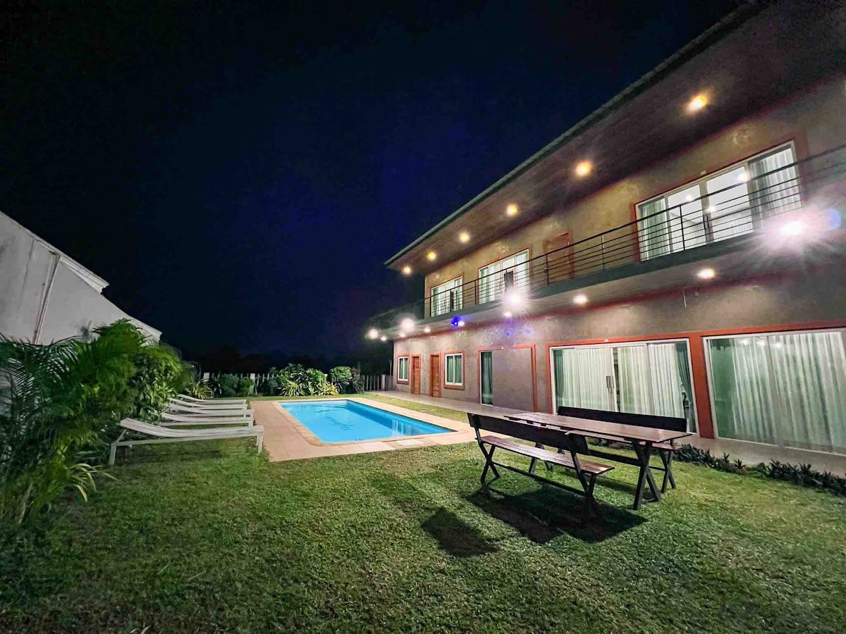 A private villa is illuminated under the night sky, featuring a swimming pool surrounded by grass. Sun loungers are arranged neatly beside the pool, and a large picnic table is positioned on the lawn, offering a gathering space for guests.