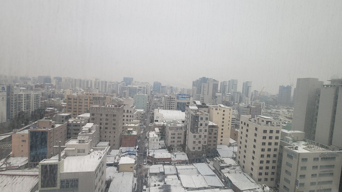 A panoramic view of a cityscape under a blanket of snow is presented. Various structures, including low-rise buildings and high-rises, can be seen. The rooftops are dusted with snow, and a soft haze covers the skyline, creating a serene winter atmosphere.