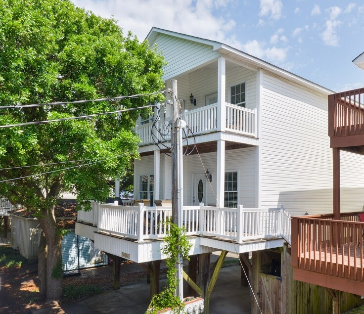 The exterior of a two-story house showcases a wraparound porch with white railings. A large tree provides shade, and nearby utility poles are visible. The house's light-colored siding contrasts with the blue sky, creating a welcoming appearance.