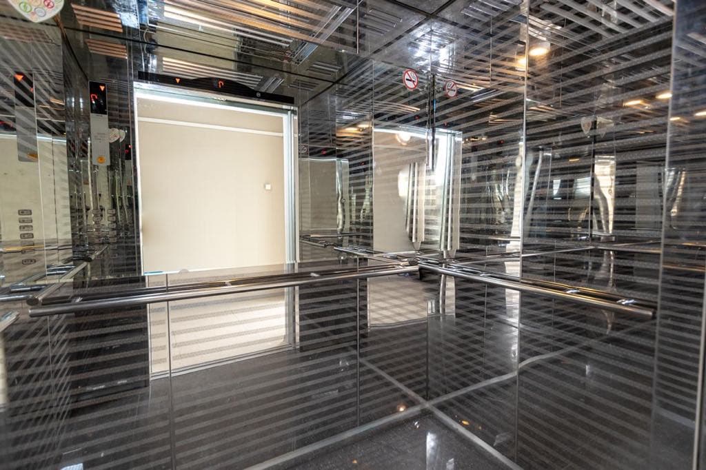 The interior of a modern elevator is shown with mirrored panels reflecting the sleek design. The floor is finished in a polished dark material, contributing to a spacious appearance. Light enters from an opening, creating a bright environment within the elevator space.