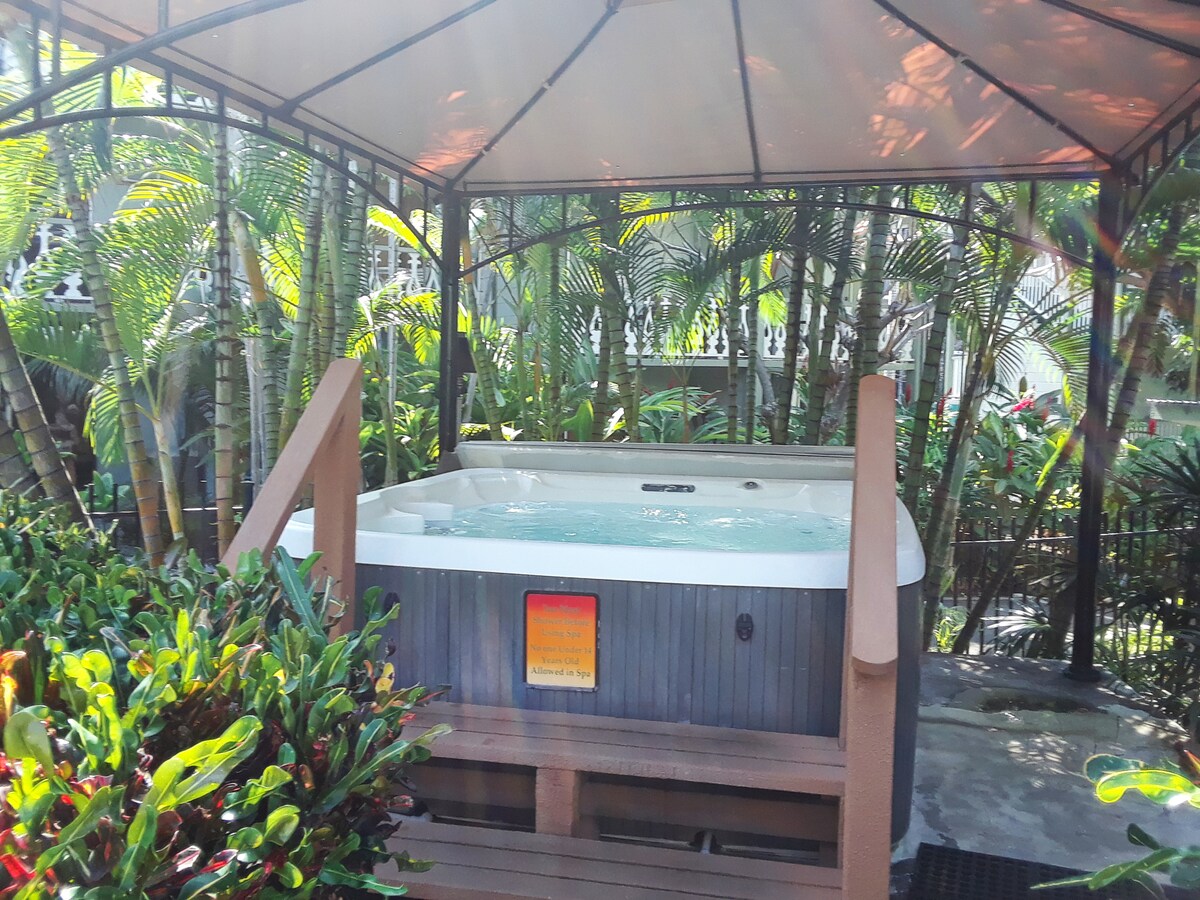 A private hot tub is enclosed within a gazebo, surrounded by lush tropical greenery and vibrant foliage. The inviting water is gently rippling, reflecting soft sunlight. A safety sign is visible near the edge, indicating rules for use.