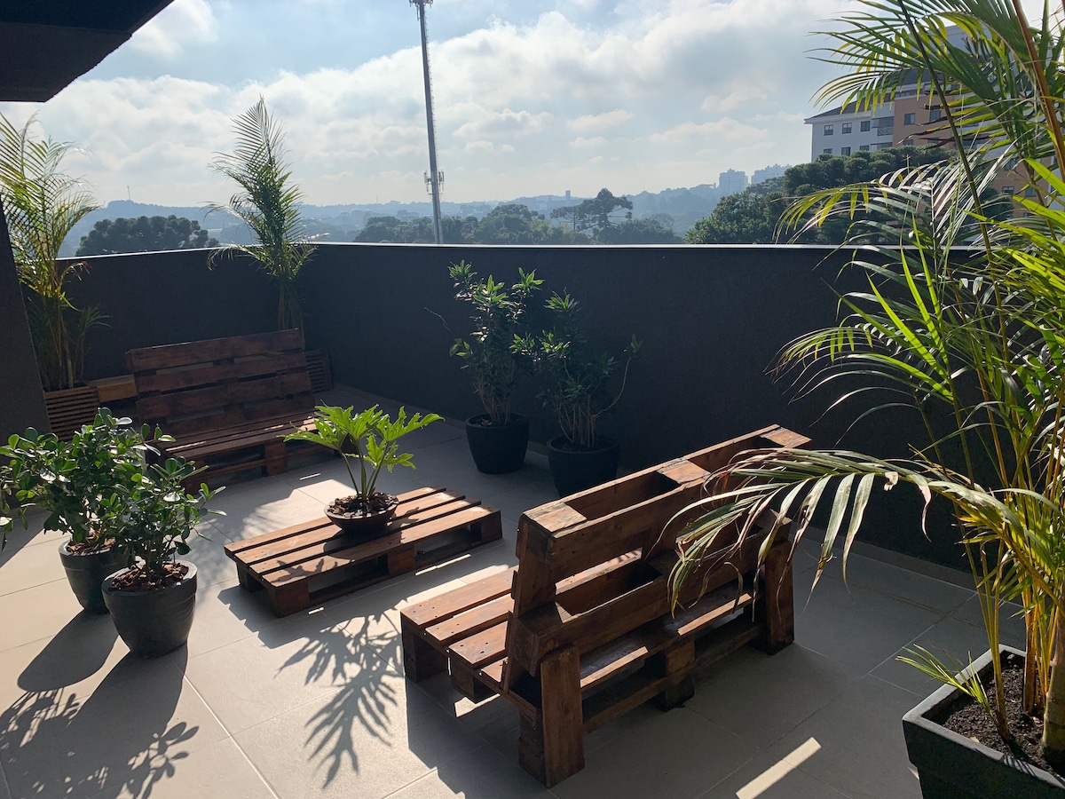 An outdoor terrace showcases wooden seating arrangements surrounded by potted plants. Bright sunlight filters through the greenery, providing a serene atmosphere. The view in the background includes distant buildings and trees, contributing to a peaceful outdoor retreat.
