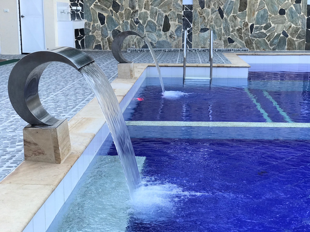 A modern swimming pool is featured, showcasing a deep blue surface and stone accents. Two sleek water features cascade into the pool, creating gentle ripples. The surrounding area consists of textured tiles and stone walls, enhancing the pool's contemporary design.