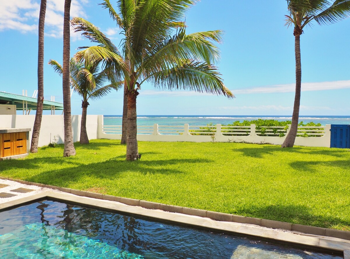 The image features a serene view of a lush green lawn bordered by gently swaying palm trees, with the ocean visible in the background. A sparkling pool lies in the foreground, reflecting the vibrant blue sky and creating a peaceful atmosphere.