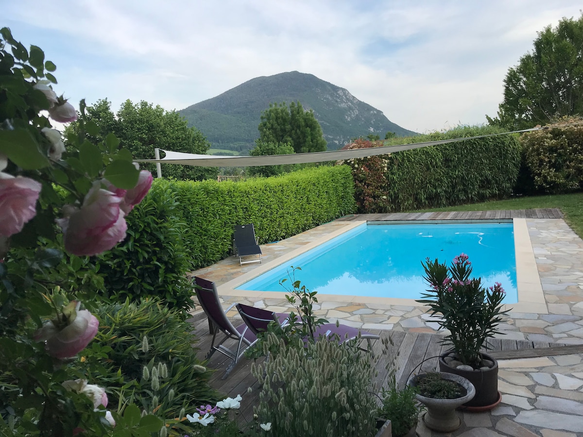 A private pool is framed by a stone patio and surrounded by lush greenery. Lounge chairs are positioned nearby, while flowering plants add color to the scene. In the background, a gentle mountain slope creates a serene view under a partly cloudy sky.