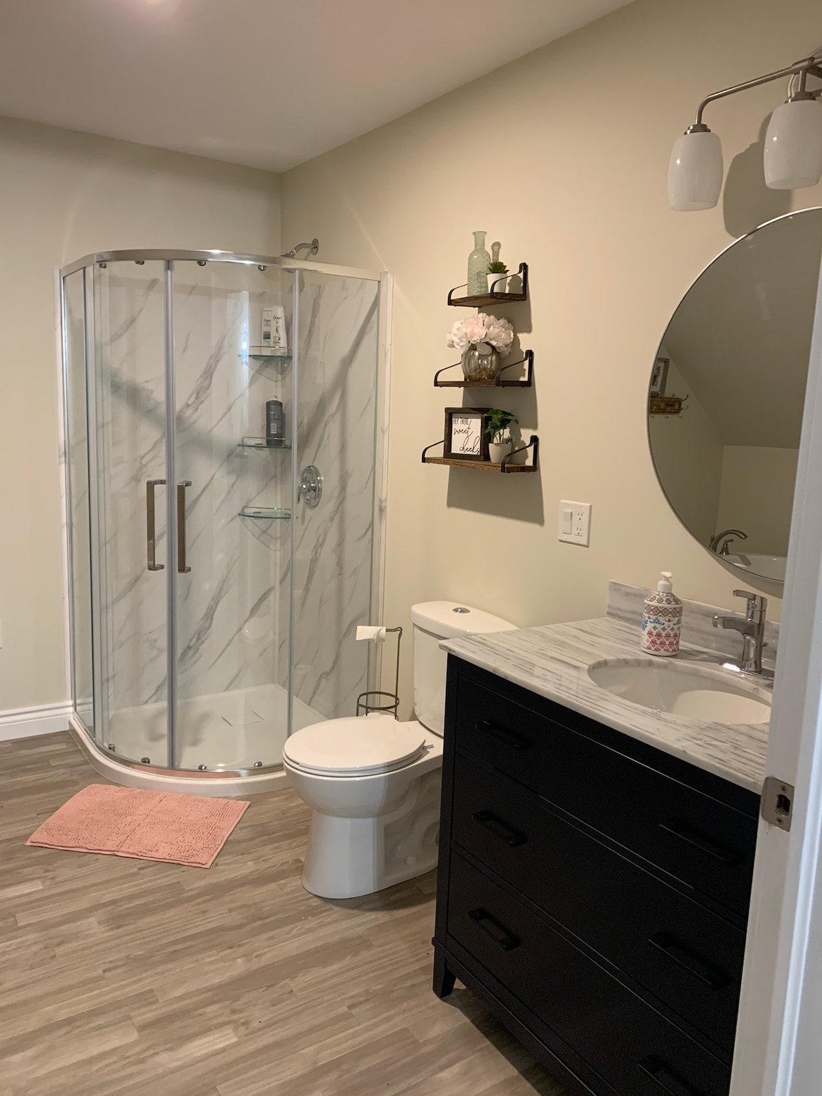 The bathroom features a modern design with a glass shower enclosure and polished marble walls. A round mirror is mounted above a stylish sink with a dark vanity. Additional decor includes wall shelves displaying potted plants and toiletries, complemented by a soft pink bath mat.