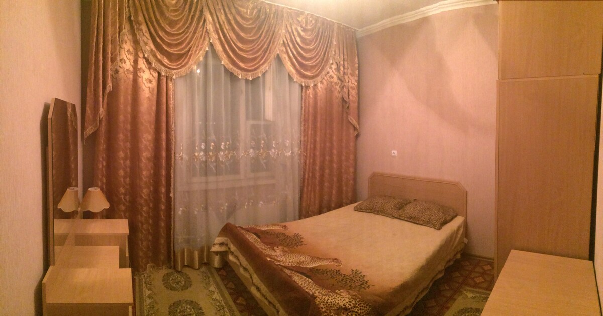 A softly lit bedroom features a spacious bed with decorative bedding. Elegant curtains frame the window, while wooden nightstands support lamps on either side. Light-colored walls and a patterned rug add warmth to the inviting space.