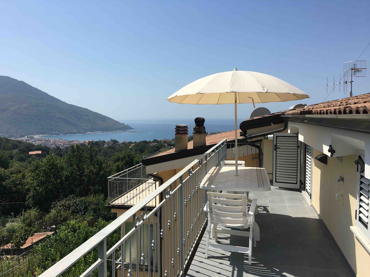 A spacious terrace offers a panoramic view of the coastline and mountains. A large white umbrella provides shade over a dining table and chairs. The area features tiled flooring and a railing that enhances the open feel, inviting relaxation and enjoyment of the natural surroundings.