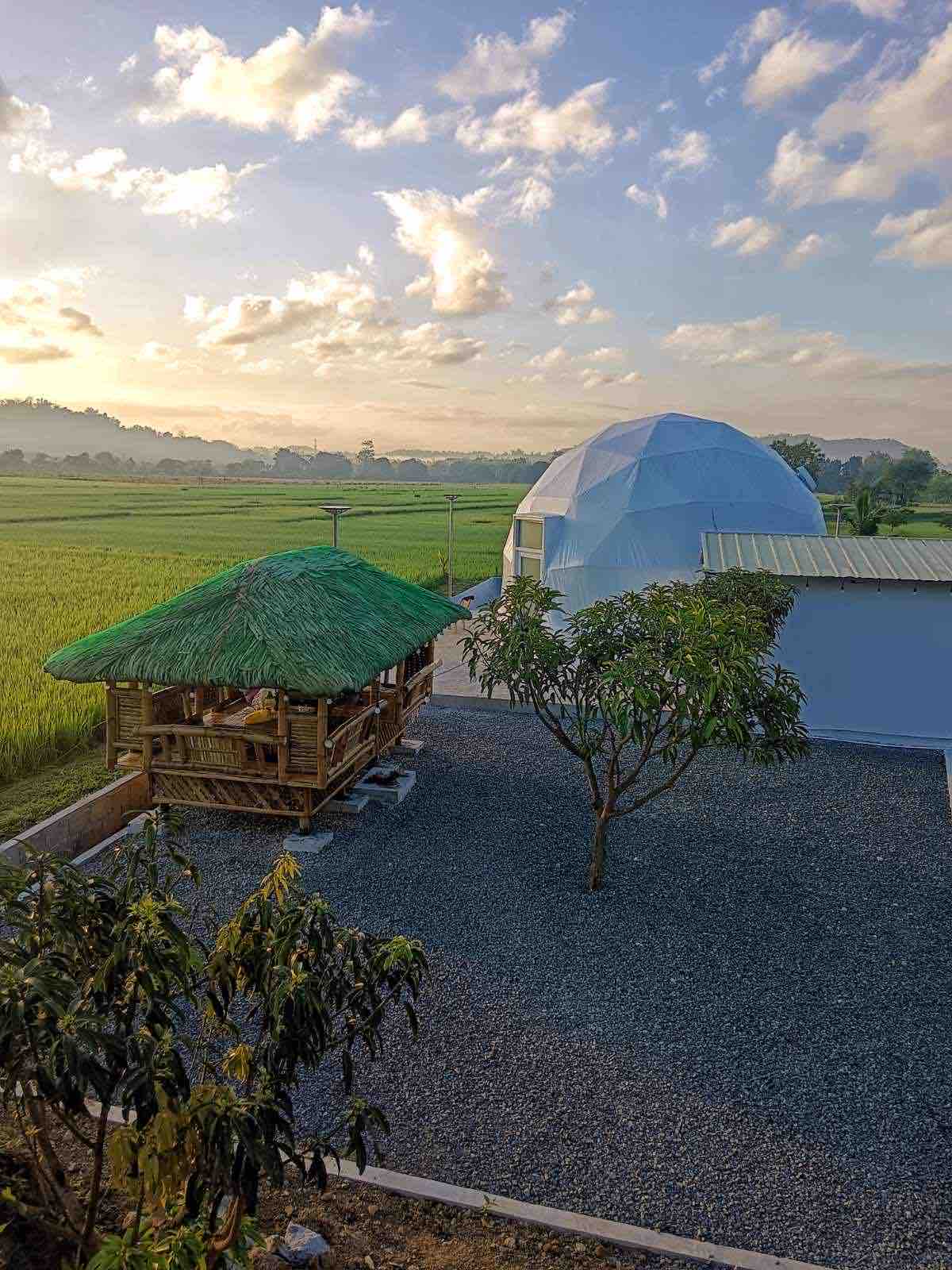 A spacious luxury dome is surrounded by a gravel area, providing a contrast to the lush green rice fields in the distance. Adjacent, a large kubo with a thatched roof offers a shaded space, adding cultural charm to the serene landscape.