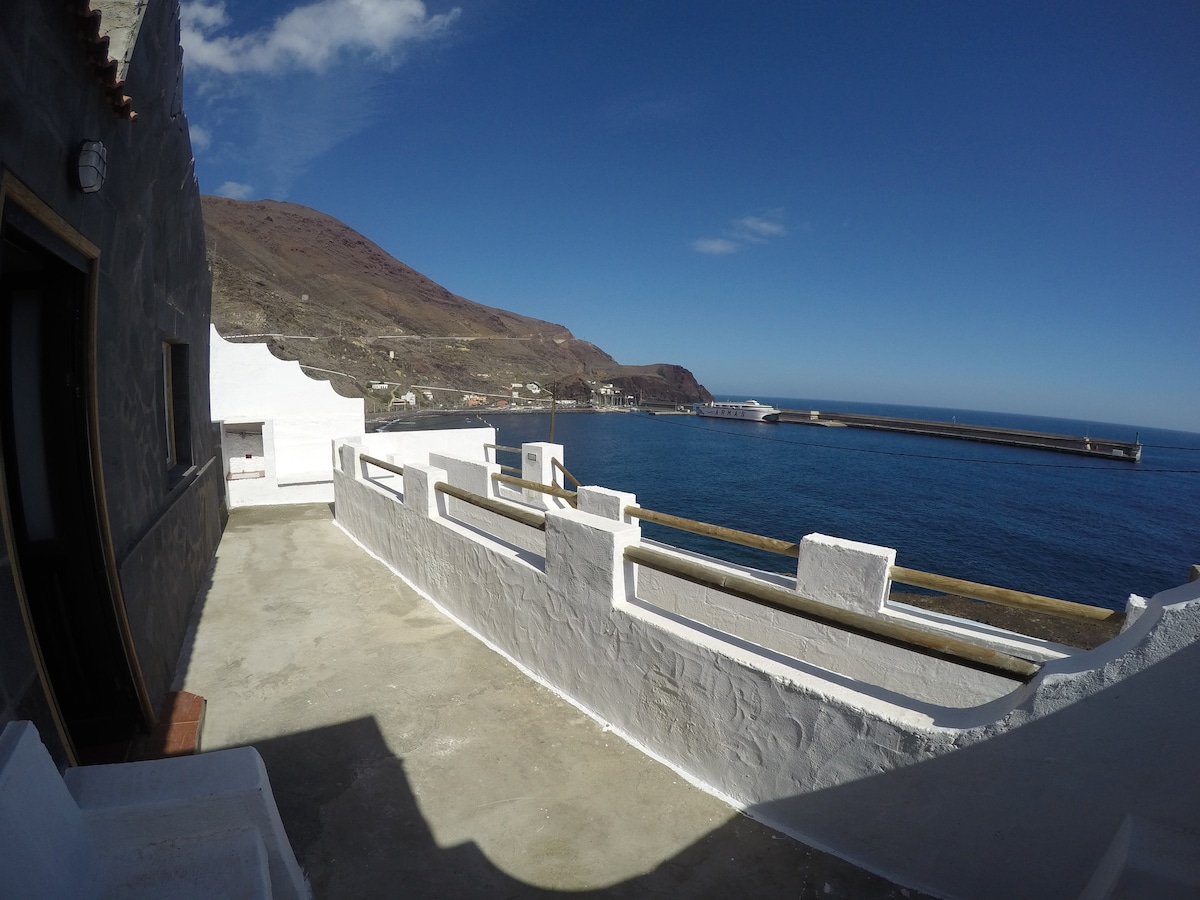 A spacious terrace offers views of the ocean and surrounding cliffs. The white walls contrast with the blue sky and water. Nearby, the harbor is visible, enhancing the coastal ambiance. The area features a smooth concrete floor, suitable for outdoor relaxation.