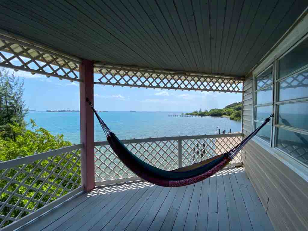 A comfortable hammock is suspended on a spacious balcony, offering views of the tranquil waterfront. The lattice design of the balcony creates a sense of openness, with sunlight filtering through, highlighting the surrounding greenery and calm water.