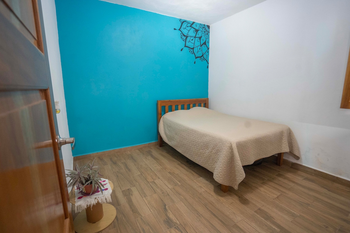 A simple sleeping area features a wooden bed with a light beige blanket. One wall is painted in a calming blue, complemented by natural wood flooring. A small decorative plant rests on a pedestal near the bed, adding a touch of greenery to the space.