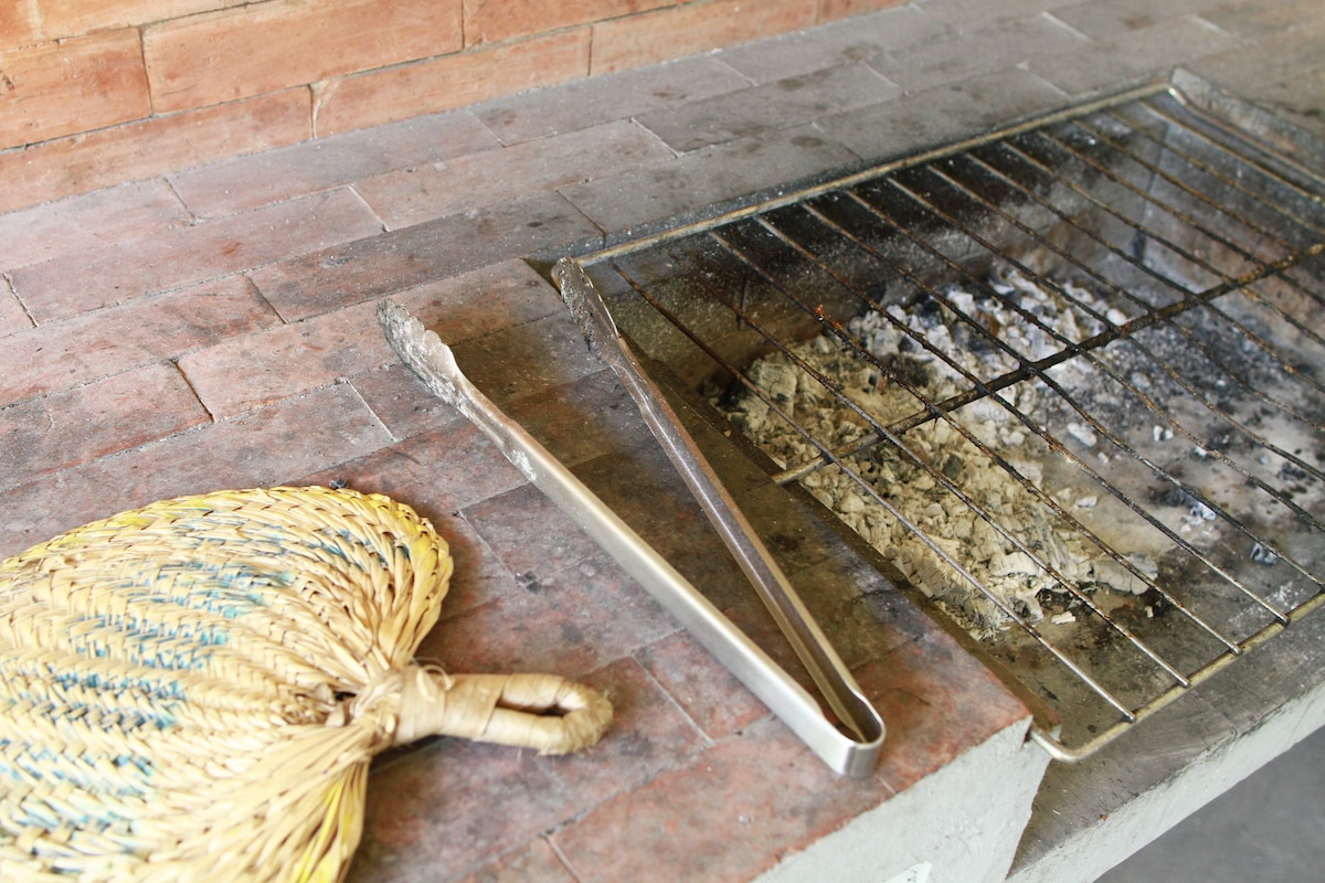 An outdoor grilling area features a metal grill with some remnants of ash and charcoal, positioned over a stone countertop. A woven fan rests nearby, suggesting a space designed for communal cooking and enjoyment in a relaxed setting.