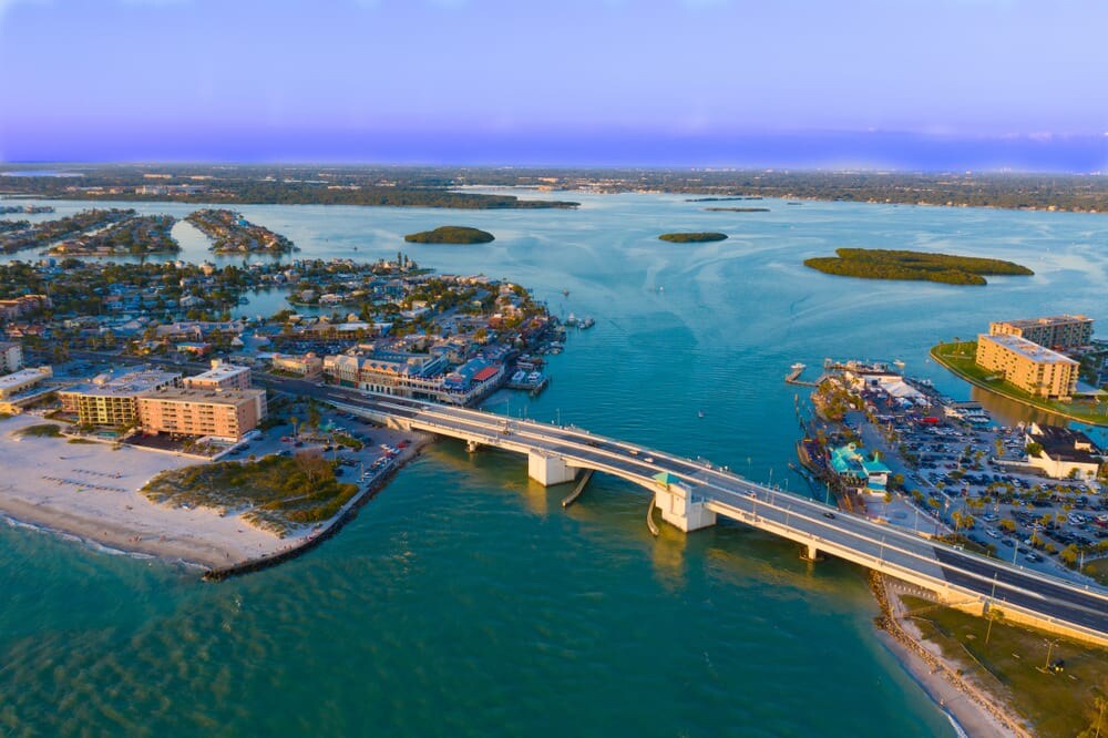An aerial view captures the vibrant waterfront community, with sandy beaches and calm waters. The bridge spans the water, connecting residential and commercial areas, while small islands are visible in the distance. The scene showcases a harmonious blend of nature and urban life.
