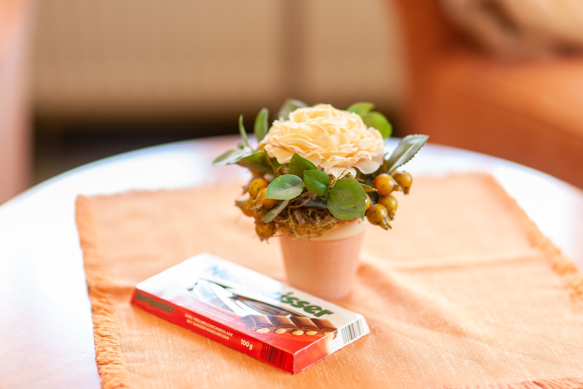 A small potted flower arrangement with a cream-colored rose and green foliage is displayed on a round table. Next to it, a chocolate bar is placed, with a light orange tablecloth providing a warm base.