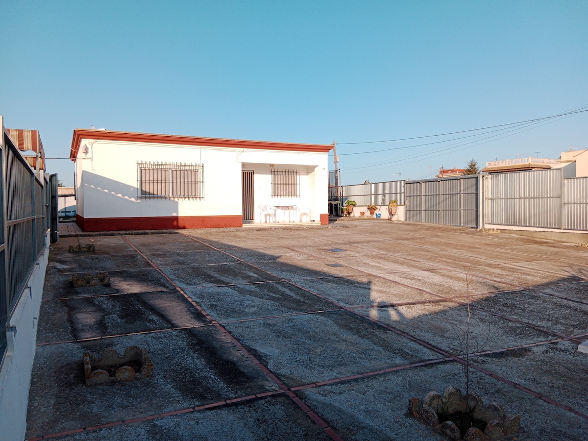 A spacious outdoor area is shown, featuring an expansive concrete surface with defined sections. In the background, a single-story white building with large windows is visible. Outdoor furniture is placed on the left, and a fenced perimeter provides privacy.