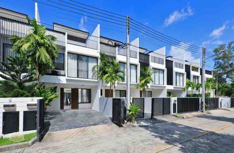 A modern townhouse facade is showcased, featuring a series of large windows and distinctive architectural lines. Lush greenery flanks the entrance, complementing the clean design. The paved driveway leads to the main entrance, which is framed by a decorative gate and fencing.