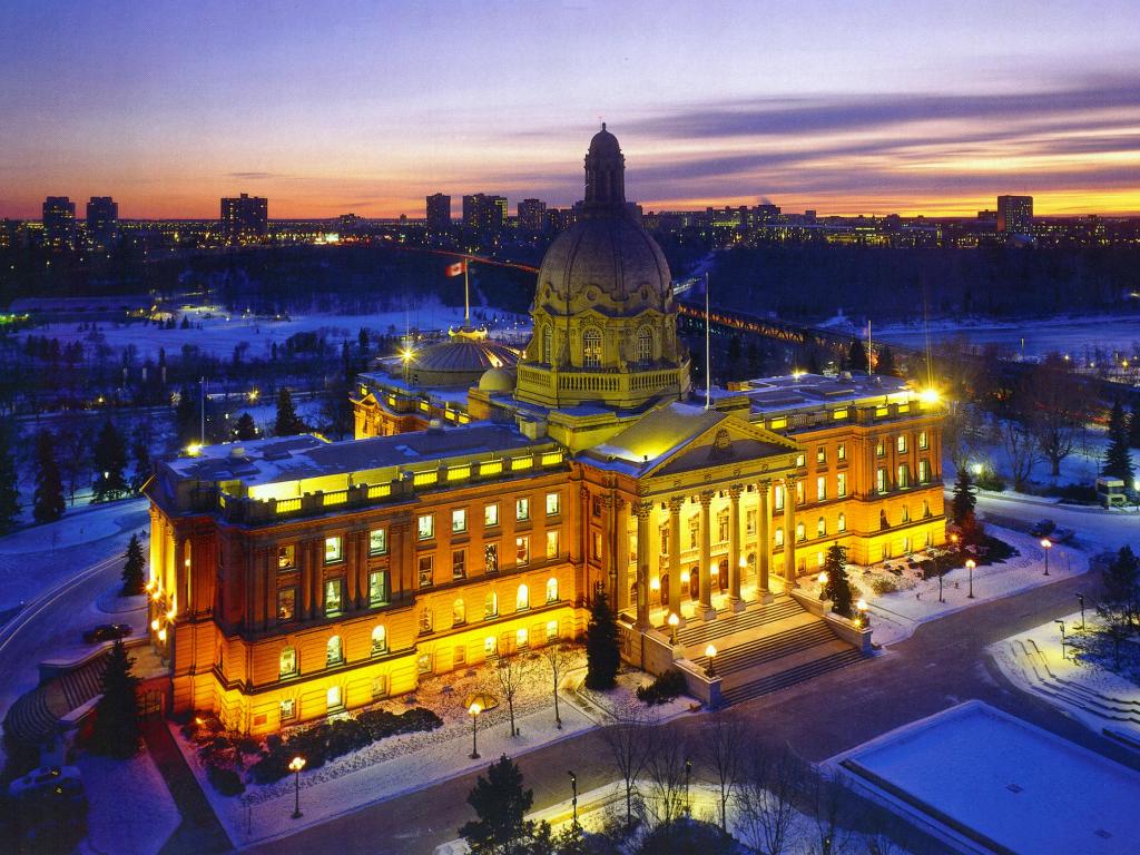 A grand historic building is illuminated at dusk, showcasing its intricate architecture. Snow blankets the grounds, while the city skyline glows in the background. Streetlights cast a warm glow on the paths, creating a serene winter landscape.