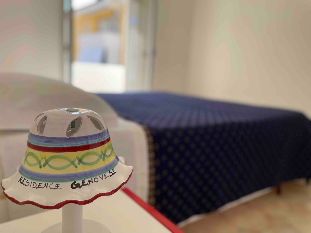 A lamp with a colorful design is positioned on a table, providing quaint charm. In the background, a spacious bed is dressed in a deep blue coverlet featuring a subtle pattern, creating a serene and comfortable sleeping area.