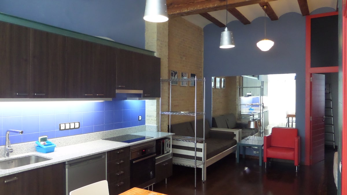 A modern kitchen area features dark cabinetry and a contrasting blue backsplash. Equipped with essential appliances, the space flows into a cozy lounge with a gray sofa. Exposed wooden beams and industrial light fixtures add character to the loft’s design.